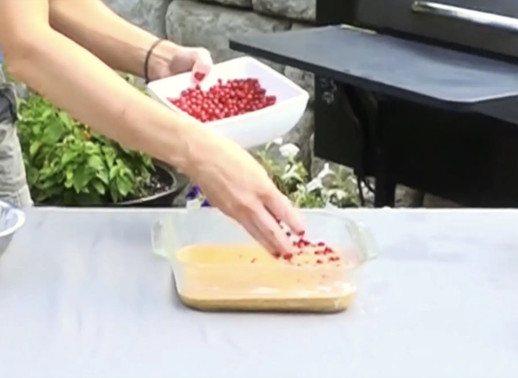 A hand scattering autumn berries over batter in a glass baking dish.