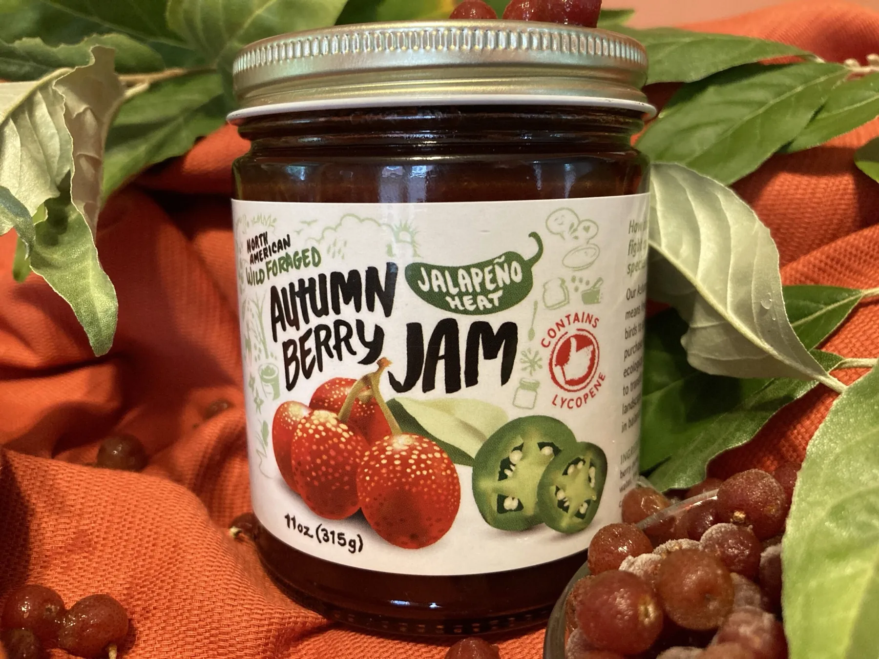 A jar of Autumn Berry Jam, jalapeño heat, among leaves and berries on orange cloth.